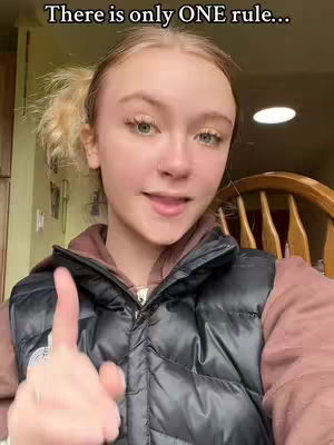 A close-up shot of a young woman with light skin, fair hair styled in two side ponytails, and a black vest over a pinkish-brown top. She is pointing her index finger towards the viewer, with a subtle smile and her tongue slightly out. The background includes hints of a kitchen setting.
