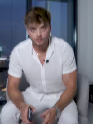 A man in a white shirt and pants sits in a chair. He is looking directly at the camera and holding a black phone-like object.