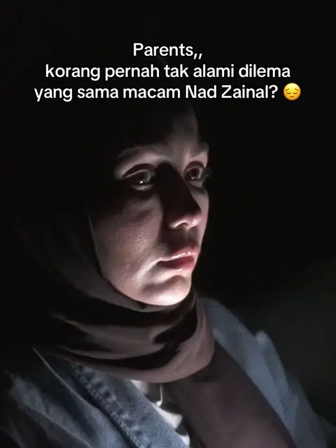 A close-up shot focuses on a woman's face, illuminated from below, suggesting a low-light setting; she wears a hijab, making her the subject of the frame and her expression suggests a sense of concern or contemplation.