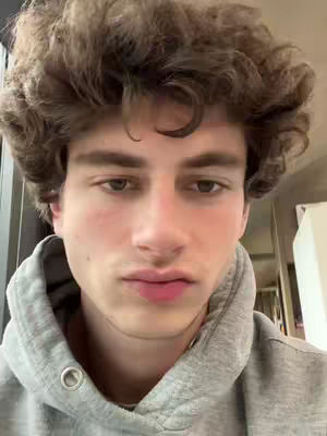 A close-up shot features a young man with light skin and brown curly hair, wearing a gray hooded sweatshirt. His expression is neutral with slightly pursed lips.