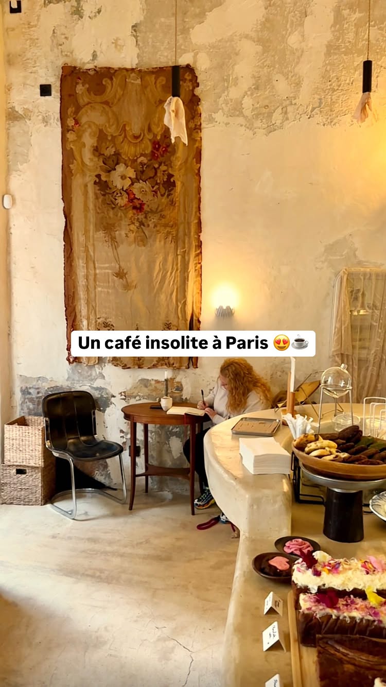 The image shows the interior of a cafe with a warm, inviting aesthetic. A person with curly blonde hair is sitting at a small wooden table in the foreground, writing in a notebook. The cafe has a rustic feel, with a large, ornate tapestry hanging on the wall behind the person. The walls are textured with visible imperfections, and there is a display of pastries and cakes on a counter in the right foreground. The lighting is soft, with a warm glow emanating from lights in the background. The general composition is a balanced shot, with the person in the center and the cafe setting creating a sense of place.