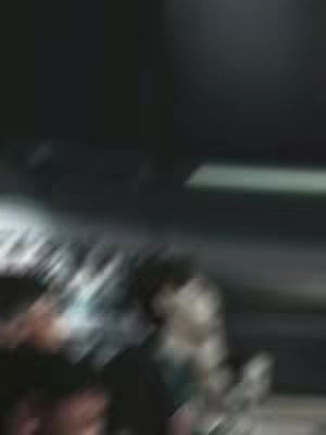 The image is heavily blurred and dark, seemingly featuring a figure in the lower portion of the frame. The background is indistinct, with only vague shapes and shadows suggesting a possible interior setting. The overall impression is one of motion blur and lack of detail.