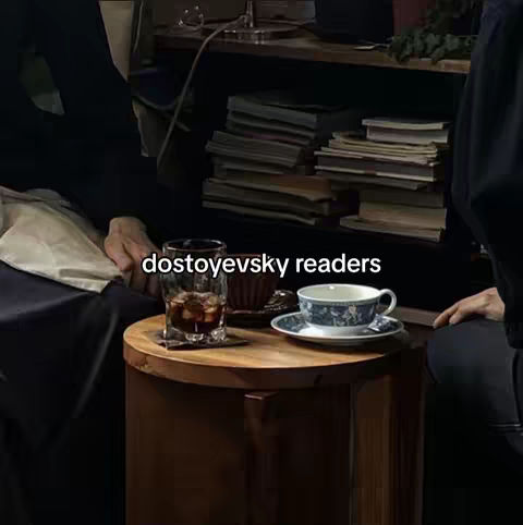 A dimly lit scene shows a round wooden table with drinks and tea things. A glass with a dark drink in it sits on a square coaster, and a blue and white teacup and saucer are to the right, along with a small, dark brown cup appearing to be the holder for teabags. A stack of books and a shelf can be seen in the background, with parts of people are in shot; one is in dark clothing and the other is wearing a light color.