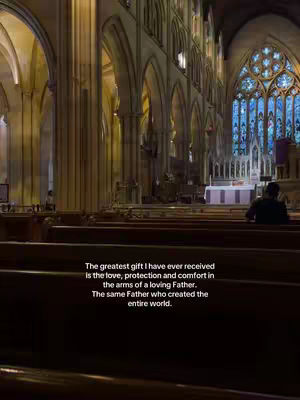 An interior shot of a church with high, arched ceilings and stained-glass windows. Wooden pews line the floor, and a person is seated in the pews away from the camera.