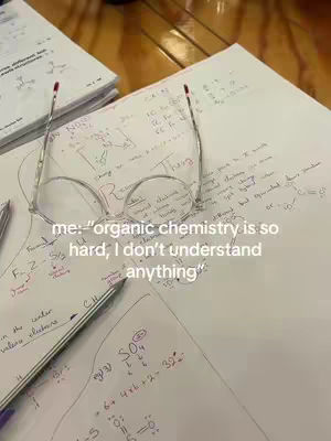 The image is a close up of a study setting. At the center are clear-framed glasses. Underneath are sheets of paper filled with handwritten chemistry equations and notes; a pen is also present, along with a stack of papers in the upper left corner. The background has a wooden surface.
