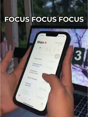 A person holding a phone with a to-do list on the screen, captured in front of a desk and window. The screen displays the calendar for Monday March 24, with a red dot indicating the current time, listing several tasks like 'Read a book', and other appointments on the list. The phone is held by hands with visible nails. Background includes a laptop with a faint background and a glimpse of foliage through a window on the left.