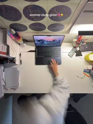 An overhead shot shows a study space. The primary focus is a laptop on a white desk, with a hand typing. The background is blurred, but suggests a workspace with study materials and a poster.