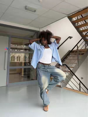 A full-length shot of a person in a modern indoor space. The person is standing with one leg bent, arms raised with hands on their head, and their body rotated towards the camera. They are wearing a blue and white striped button-down shirt over a white tank top, light-washed, baggy jeans, and tan colored shoes. The background shows a white ceiling with fluorescent lights, glass doors, the beginning of a staircase, and a gray floor.