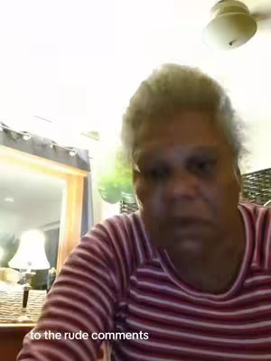 A woman with short gray hair wearing a striped shirt is looking at the camera with a serious expression. The background includes a mirror and some household items.