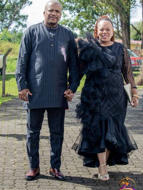 A well-lit outdoor shot features a couple, a man and a woman, holding hands and walking towards the camera. The man is on the left, wearing a dark, vertically striped suit, burgundy shoes, and a pinstriped shirt. The woman, to his right, is in a full-length black dress with decorative ruffles and a tulle skirt, paired with gold heels. They are positioned on a brick-patterned paved area, with blurred greenery and a glimpse of a red car in the background. The overall impression is one of celebration and elegance, likely taken during daylight.