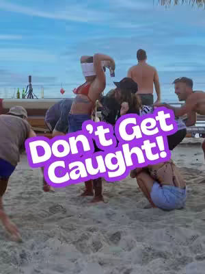 A group of people on a beach, playing a game. A person is blindfolded and holding an object, possibly a camera. A text overlay reads 'Don't Get Caught!'. The scene is lit by daylight with a beach and ocean backdrop.