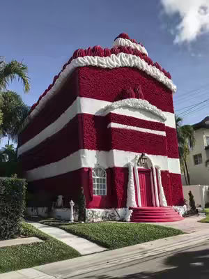 A house designed to look like a giant red velvet cake, with red and white striped walls, a peaked roof with white frosting details, and a pink door. The house is set on a well-manicured lawn with palm trees and a bright blue sky.