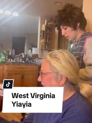 A candid vertical shot showing an older woman and a younger person in a kitchen setting. The older woman, with light-colored hair and glasses, is in the foreground, facing left. The younger person, with curly hair, is behind her, looking off to the side. A white rectangular text overlay near the bottom reads "West Virginia Yiayia," indicating the content's theme.