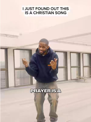 The image shows a man in a dark blue hoodie and grey pants standing slightly on the right side of the frame with both hands up in the air making a 'hang loose' gesture. The man seems to be in an open space with windows in the background and a low ceiling with what appears to be a light fixture. There's text at the top and the bottom of the image. The overall tone of the image appears to be positive.