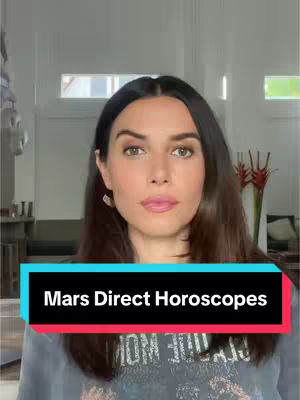 A woman with dark hair and light skin is centered in front of a camera. She is wearing earrings and a grey t-shirt with the words 'OM 333' visible. A black rectangle with red outlines features the text 'Mars Direct Horoscopes'. The background includes a brightly lit indoor space with windows, a sofa, and red decorative flowers.