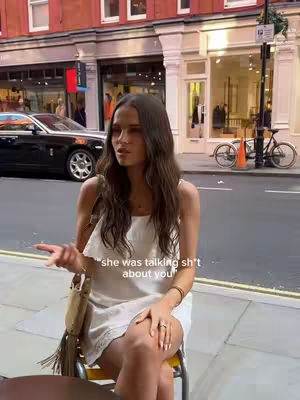 A woman is seated on a chair outside next to a street. She has long brown hair and is wearing a white dress. In the background, there are shops and a car. Text overlay reads, "she was talking sh*t about you."
