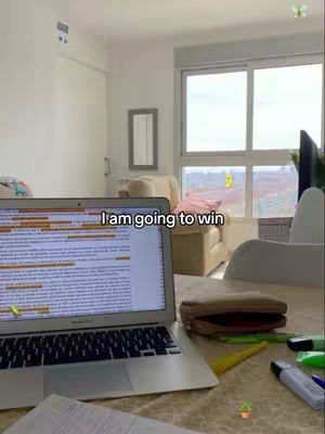 The image shows a laptop open on a table with a window and some furniture in the background. The laptop screen displays text, with some parts highlighted. A handwritten note and pens are nearby. The words 'I am going to win' are overlaid on the image.