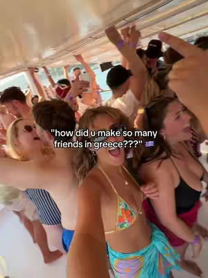 A group of young adults on a boat party in Greece. Text overlayed reads, \"how did u make so many friends in greece???\" The person filming is a young woman in a bikini, surrounded by other people dancing and interacting.