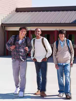 Three young men are walking outdoors on a sunny day, likely on a school campus, each with a backpack. They are smiling and appear to be casually conversing. The background includes a building with an awning and red accents.