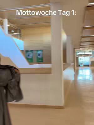 The image shows a hallway with beige walls and flooring. A person, partially visible on the left, appears to be walking towards the camera. The hallway extends into the distance, with a staircase and other architectural features included. The text overlay at the top reads: 'Mottowoche Tag 1:'