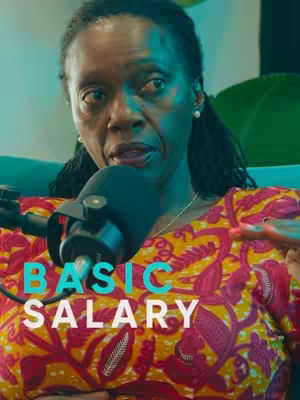 A woman with dark hair and a microphone is the central focus. The text, "BASIC SALARY," is superimposed over her torso. She wears a bright, patterned top in shades of yellow and red. The background appears to be a blurred, indoor setting.