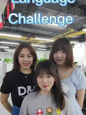 A group of three women posing for a photo. The central figure is wearing a gray t-shirt, while the other two are wearing black and blue tops. Above, the text 'Language Challenge' is written with colorful gradients.