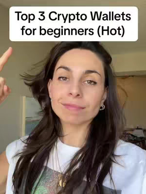 A woman with long dark hair smiles slightly, pointing a finger upwards. Above her is a white banner with black text that reads "Top 3 Crypto Wallets for beginners (Hot)". The woman is wearing a white t-shirt and a necklace. The background slightly blurred, appears to be a domestic setting.
