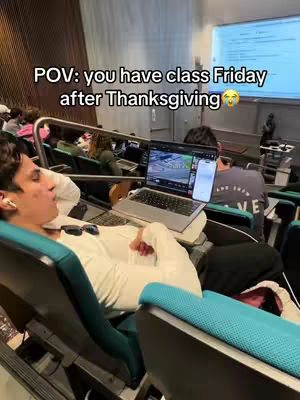A person lies in a chair in a lecture hall, appearing asleep. A laptop and phone rest on the attached desk.