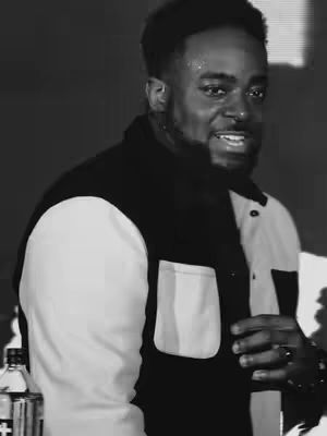 A black and white shot of a man smiling. He is wearing a black and white collared shirt and has a focused gaze. The background is a dark, blurred setting. There's a bottle of a liquid in the lower left.
