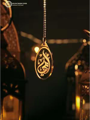 A close-up shot featuring a gold pendant hanging from a delicate chain. The pendant has an oval shape and features elegant Arabic calligraphy. It hangs against a dark, blurred background that creates a sense of depth and highlights the pendant's design. The setup suggests a product presentation, possibly for a jewelry collection.