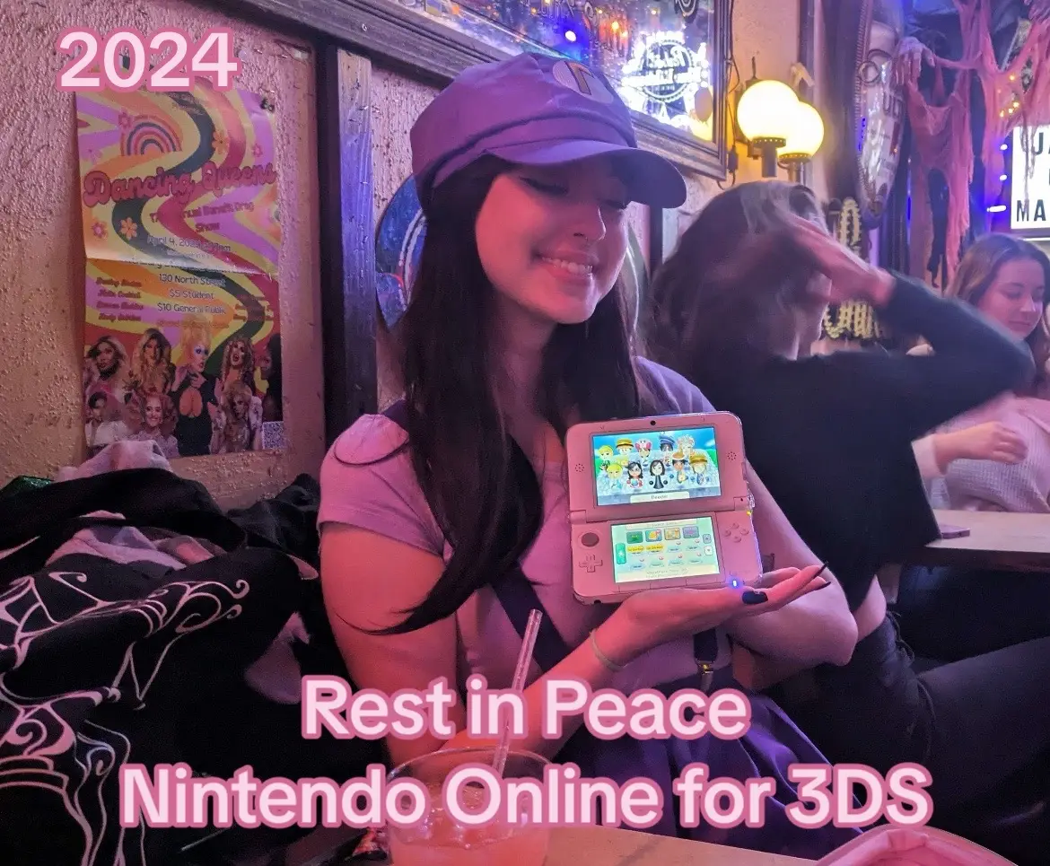 A selfie-style photo taken in a bar setting. The primary focus is on a young woman with long dark hair wearing a purple hat and shirt, holding a pink Nintendo 3DS open, facing towards the camera. The screen displays a game with colorful characters. Behind her, other people are visible, and the background includes a colorful poster and bar decorations.  The overall composition gives a nostalgic and personal vibe.