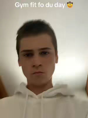 A selfie-style shot of a young man from the chest up, set inside. He's wearing a white hoodie, the hood down, and his expression is neutral with a slight frown. The background is a plain, light-colored wall. Text overlay at the top reads 'Gym fit fo du day' with an emoji, suggesting a gym or fitness context.
