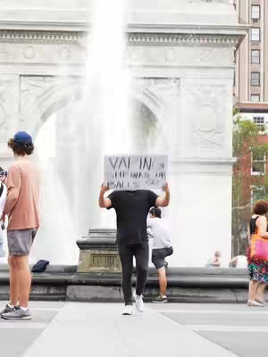 An outdoor shot featuring a fountain in the background and several people in the scene. A person in the foreground is holding a sign with text written on it, while other individuals are visible in the distance. The sky is bright, and the image has an overall daytime setting.