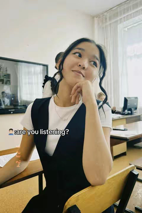 A young woman with dark hair is seated at a desk in a brightly lit classroom. She is wearing a white top and a black vest, and is looking off to the side with a thoughtful expression, her hand near her chin. A hint of hair from what could be a second person in the background adds depth, and the image has the casual feel of a candid shot captured in a learning environment. The lighting is natural and the color palette leans towards cool tones.