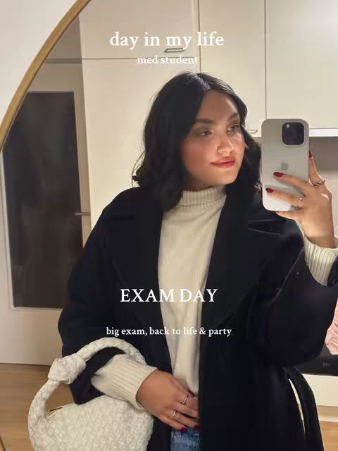 A woman is holding a phone up to record herself, while posing in a mirror. She is wearing a black coat over a white sweater, and a white purse is in view. The text overlay says "day in my life med student" and "EXAM DAY big exam, back to life & party"