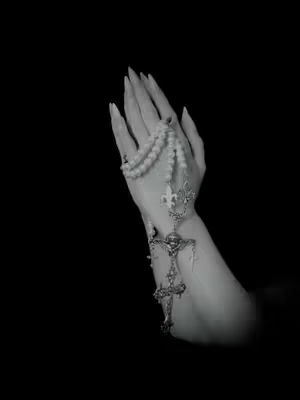A black and white image reveals a hand, adorned with a rosary, set against a stark black background. The hand is positioned with the palm open, fingers gently curved as if in prayer. A rosary with gleaming beads, accented by a detailed cross and decorative embellishments, drapes from the hand. The lighting is focused, highlighting the hand and the rosary, while the deep black background maximizes the contrast.