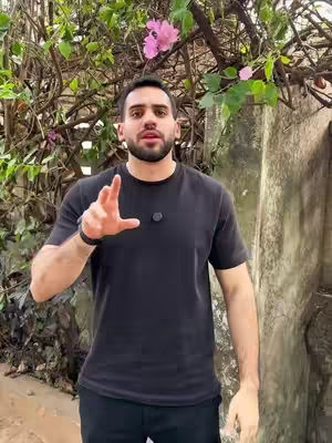A man in a black t-shirt is standing outside in front of some green foliage and a stone wall. He is pointing his hand out towards the viewer. In the background is some green foliage and light pink flowers.