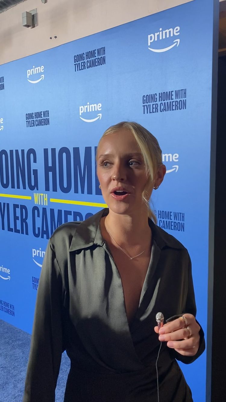A woman with blonde hair in a ponytail is speaking into a microphone while standing in front of a blue backdrop. The backdrop has the Amazon Prime logo repeated multiple times, along with text that reads "Going Home with Tyler Cameron". The woman is wearing a dark green satin top and a delicate necklace and rings. The upper lighting adds to the professional setting, suggesting an interview or a promotional event.