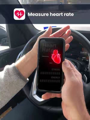 The image shows a person holding a smartphone displaying a heart rate monitor app. The person is in a car, with the steering wheel visible. The app's interface shows a red heart graphic and data relating to heart rate, stress, and energy levels. A text overlay at the top reads, "Measure heart rate."