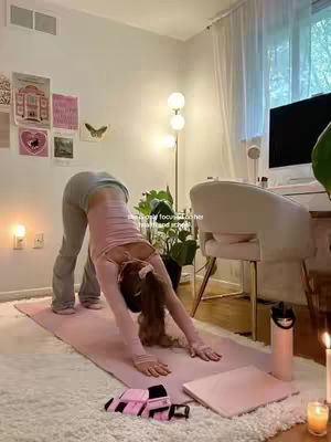 A woman in exercise clothes is in a yoga pose (downward dog), hands and feet on a pink mat, with a light-filled room as a background. The room's decor has posters with some decorative items. A laptop and water bottle are on the floor next to her.