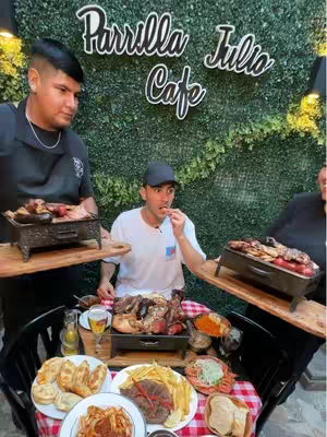 A group of people are gathered around a table laden with various grilled meats, sides, and drinks. A man in a white shirt is eating a piece of food with an animated expression, positioned in the center of the image. Two individuals, one on each side of the central figure, hold black trays with grilled items, seemingly serving the main course. A variety of other dishes and items such as fries, salads, and glasses of beer are spread on and around the table, accompanied by a checkered red and white table cloth. The background features a green wall installation, topped by a sign in a handwritten font.