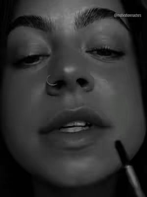A close-up, black and white portrait of a person, focusing on their face. The person is looking down with a slightly open mouth, a small ring in their nostril, and the lower portion of their face is the main focus. An applicator is held near their mouth, suggesting they are applying makeup. The overall composition is striking, utilizing stark contrasts and a shallow depth of field.
