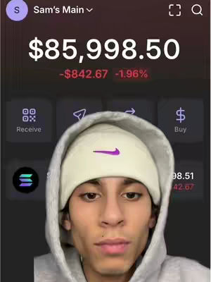A close-up shot of a young man wearing a gray hoodie and a white beanie with a purple Nike swoosh. The background shows a mobile app interface with a large dollar amount ($85,998.50) displayed prominently at the top, along with financial details indicating a loss of 1.96%. Below the man are icons for 'Receive' and 'Buy'. The overall composition suggests content related to finance or cryptocurrency.