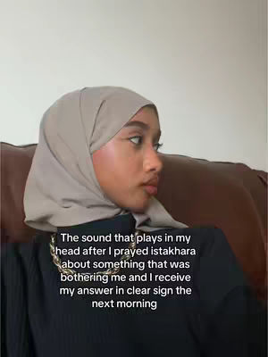A woman in a hijab sits in an indoor setting, likely on a couch. The image is a close-up of her face and shoulders.