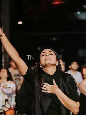 A woman in a dark shirt and black vest is raising her right arm in worship in front of a crowd of people. Text overlayed reads "The".