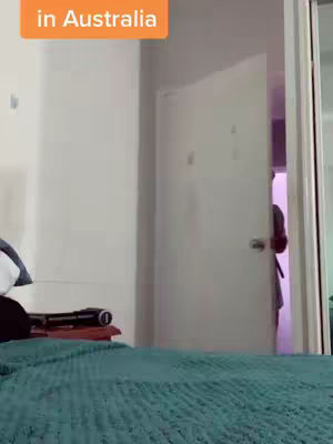 The cover image shows a room with a bed in the foreground covered with a teal bedspread. Behind the bed, a white door is ajar, revealing a person wearing a robe in a room with a purple hue. The image has an orange text overlay at the top, likely the video's title.