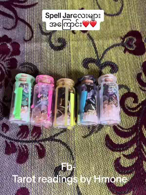 The image shows five small, clear jars filled with various colorful items. The jars are arranged horizontally against a patterned background with floral and leaf motifs in shades of maroon, green, and yellow. Each jar seems to contain a different assortment of materials, including what appears to be small crystals, colorful beads, and other decorative elements. The jars are sealed with lids and have what looks like neon colored wax drips on top. Above the jars, the text overlay reads 'Spell Jar' in a white box. Below, smaller text indicates a Facebook account.