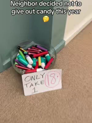 The image shows a clear glass bowl on the ground beside a wall; the bowl contains a collection of colorful tubes, potentially candy or small treats. A white rectangular sign is in front of the bowl, with handwritten text indicating, “ONLY TAKE 1” and a circled number '18.' The setting appears to be indoors, and the image suggests a Halloween or similar event.