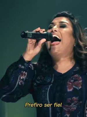 A medium shot captures a woman with dark hair and makeup, holding a microphone near her mouth and singing with great emotion. A lower-third text overlay reads 'Prefiro ser fiel'. The background is softly blurred, with a dark, concert-like lighting setup.