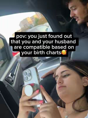 A woman is sitting in a car looking at her phone. A man is partially visible in the background. Text on the image says "pov: you just found out that you and your husband are compatible based on your birth charts" with a smiling face emoji.